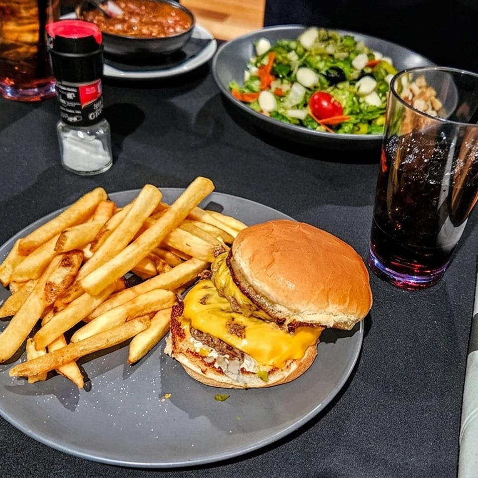 Cheeseburger with French fries, salad, cola, and condiments arranged on a dark restaurant table.